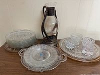 Photo showing vintage silver plated coffee carafe with ornate warmer stand surrounded by various sizes of clear cut glass plates and bowls arranged on a white surface against a white wall.