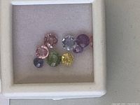 Seven assorted cut gemstones in various colors displayed in a small white display box.