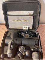 Full view of Wahme W1 body massager and attachments inside black carrying case with mesh pocket containing document.