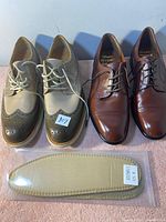 Two pairs of men's dress shoes and one pair of insoles on table. Left beige and olive green wingtip brogue shoes, right brown dress shoes, one beige insole in packaging in foreground.
