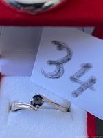Close-up photo of a silver ring with black gemstone held by a note with number 34, showing details of the prong setting and band design.