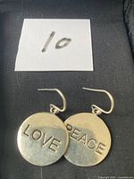 Front view of the 'LOVE' and 'PEACE' silver earrings placed side by side on a black surface with a white label marked '10'.
