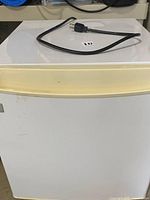 Front view of the small white mini fridge showing the yellow-toned handle and power cord on top.