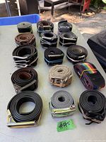 Photo showing 11 rolled belts on a table outdoors, various leather and fabric belts, buckles mostly silver-toned with some gold accents.