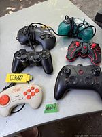Photo showing seven assorted video game controllers including Sony PlayStation and B!a$y branded controllers and adapters, all with wires or USB connectors, laid on a grey table.
