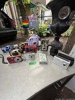 Wide view of the lot showing multiple vintage cameras including a purple Fujifilm Instax Mini instant camera, disposable Kodak cameras, box cameras with dual lenses, and a black camera lens.