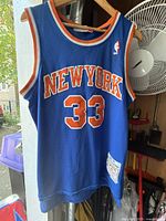 Front view of vintage blue Patrick Ewing New York Knicks NBA jersey hanging on hanger showing 'NEW YORK' and number 33 in orange with white outline, with NBA logo on shoulder.