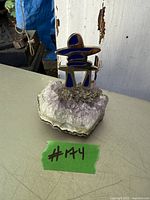 Amethyst crystal cluster with a decorative metal Inukshuk featuring blue inlays on a rough stone base shown on a table with a green lot number tag.