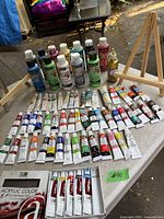 Full view of table showing multiple DecoArt acrylic paint bottles, many tubes of paints, and two mini easels.
