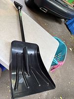 Black plastic snow shovel assembled on table next to car tire, showing the shovel blade and handle in one piece.