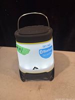 Front view of the Ozark Trail LED camping light showing its white glowing body, black top and base, and Bluetooth sticker.