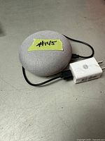 Mini Google Nest smart speaker with power adapter on a flat surface. The speaker is light gray fabric top and connected to a white power adapter.