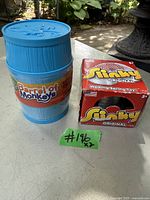 Photo showing the Original Slinky in its original red box with yellow and white branding alongside a blue Barrel of Monkeys toy container on a table outdoors.