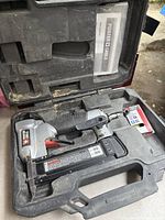 Porter Cable finish nailer in black and silver with a rectangular nail magazine, placed inside a black plastic molded carrying case with manual included in the lid compartment.