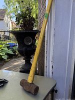 Photo of a sledge hammer leaning against a wall outdoors on a table, showing the yellow wooden handle and rusted metal hammer head.