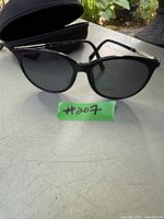 Full view of Swarovski ladies sunglasses with black frames and dark lenses along with the black protective case, placed on a gray surface.