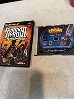 Photo showing Guitar Hero III: Legends of Rock case and Crash Bandicoot PS2 game disc on a beige surface.