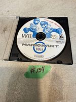 Close-up image of Mario Kart Wii game disc inside a clear DVD case. The disc shows the title and features blue artwork of Mario and Luigi on a white background.