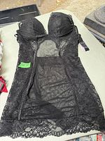 Photo displaying black lace mesh lingerie laid flat, showing padded cups, floral lace sides, mesh body, and detailed lace hem.