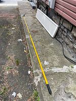 Full length view of yellow fiberglass extendible pole saw/pruner laid out on a concrete ledge and asphalt ground.