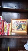 Books and framed advertisement on wooden shelf with visible titles and spines.