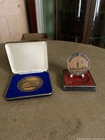Two commemorative medallions displayed side-by-side on a surface, one round within a blue velvet-lined case and one octagonal in a red-lined display case.