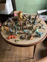 Wide view of around 30 dinosaur figurines arranged on a round table. Varied species and colors visible including Pterodactyl and Stegosaurus.