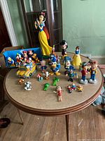 Overview of various figurines and collectibles displayed on round wooden table including Snow White large figurine, Looney Tunes characters, and other cartoon figures.