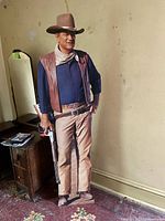 Front view of life-size John Wayne cowboy cardboard cutout in a room with wood furniture and patterned carpet.