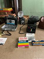 Wide view showing two vintage Polaroid cameras, a third vintage camera with flash tower, film boxes, and carrying cases on table.