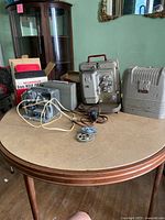 Photo of three items on round table: Keystone 8 mm projector, Academy 8 mm film editor missing front cover, Brumberger 8 mm battery-operated projector with box and reels