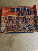 Photo of the original box with 'Mechanical Shooting Gallery' text and cartoon crowd illustration, showing wear and tear.