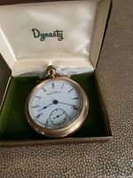 Front view of the vintage pocket watch inside a green Dynasty branded box showing the white enamel dial, Roman numerals, and small seconds subdial.