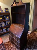 Full view of vintage wooden secretary with open shelving on top, hinged slant front panel closed, and three curved drawers below.