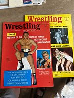 Two Wrestling Monthly magazines with cover photos of wrestlers, one prominently featuring a wrestler wearing a championship belt, the other featuring a woman and a wrestling scene on a yellow background.