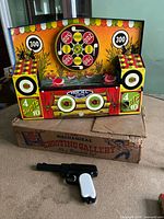 Front view of vintage metal mechanical shooting gallery toy on damaged original cardboard box with black and white target dart pistol placed in front.