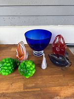 Seven assorted art glass items displayed on wooden surface against white wall, including blue bowl, red basket, green grape clusters, and various glass sculptures.