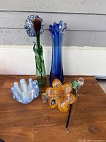 Display of all six art glass pieces including two vases, ruffled bowl, and four flower figurines arranged on wooden surface.