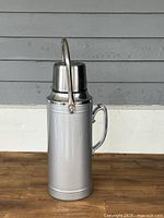 Front view of large silver metal thermos flask with handle and cup lid, placed against gray wall.
