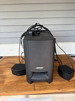 Front view of the black Bose Acoustimass subwoofer with two satellite speakers and tangled wires on a wooden surface.