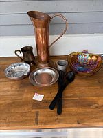 Photo showing tall copper pitcher, small dark metal cup, silver-tone plate, two black serving utensils, iridescent glass bowl, small silver-tone dish and cup on wooden surface