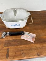 Corning Ware casserole dish in white ceramic with blue cornflower pattern, glass lid placed on top, black handled lid lifter and metal holder beneath on wooden surface.
