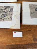 Two matted sketches showing Mickey hugging Minnie (left) and the kissing scene with “SMACK” text (right), laid on wood surface with reference card.