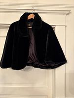 Black faux fur capelet hanging on wooden hanger showing short sleeve and wide collar.