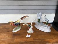 Porcelain bird figurines set including flying pheasant, mallard duck head, and pair of herons on an oval base all displayed on a wooden surface against a gray wall.