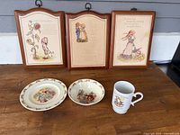 Wide view showing three framed Holly Hobby prints on wooden frames placed behind two decorative plates and one mug on wooden table.