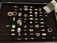 Top down view of a black jewelry tray holding 47 assorted costume rings with varied styles, colors and materials.