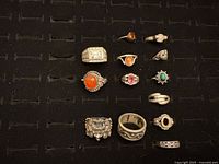 Twelve sterling silver rings placed on a black display board, showing various sizes and gemstone colors including orange, pink, green, black, and clear stones, some with ornate silver detailing.