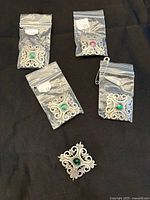 Five silver-tone filigree brooches with various colored central stones. Four are in small plastic bags, one is loose on black background.