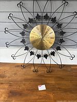 Front view of mid-century modern wall clock with black metal spokes and leaf accents, showing the gold clock face with Roman numerals.